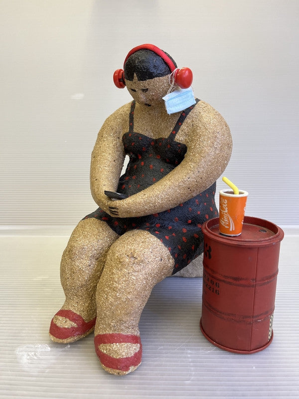 Seated sculpture of a person with red headphones, a blue mask on their arm, holding a phone. A red soda cup with a straw sits on a barrel. Playful tone.