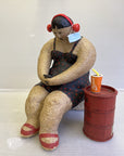 Seated sculpture of a person with red headphones, a blue mask on their arm, holding a phone. A red soda cup with a straw sits on a barrel. Playful tone.