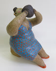 A textured sculpture of a seated figure in a blue dress with red polka dots, holding a dark round object to its mouth.