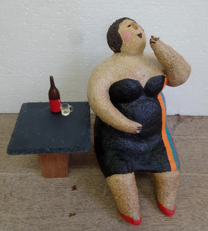 A clay figurine of a woman in a black dress eating, seated beside a small table with a beer bottle and glass on it.